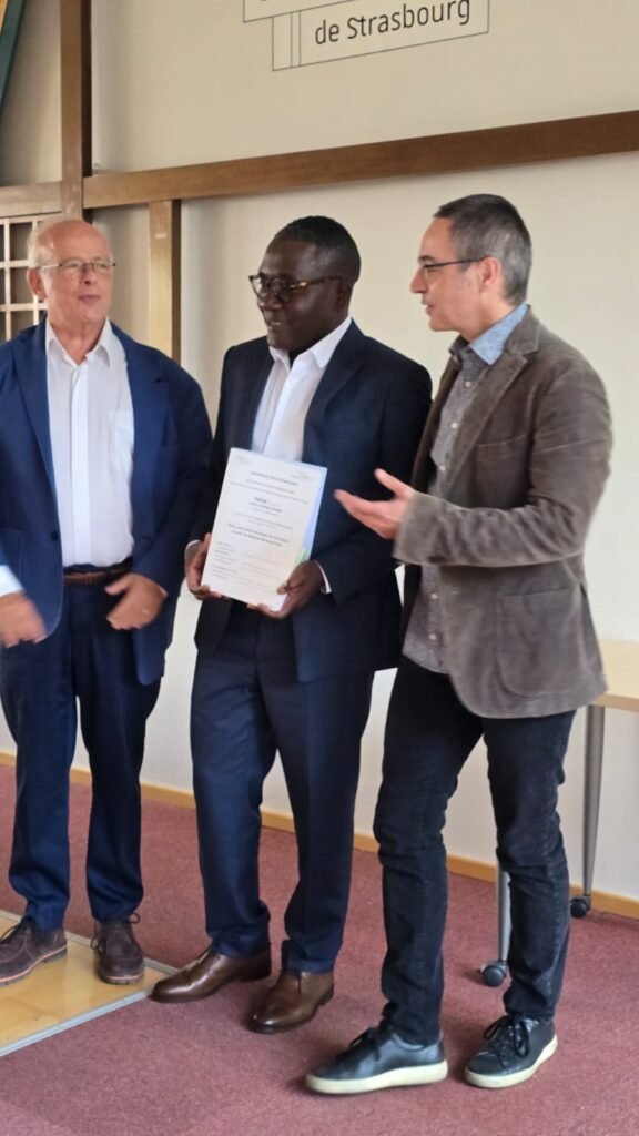 Fr Landry N’nang Ekomie smiling as he holds his thesis, flanked by two members of the jury