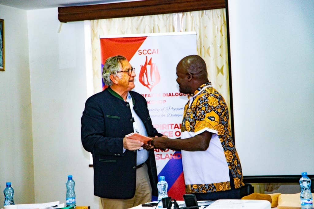 Fr. Fredrick Elima Wafula shaking hands with a guest in front of the SCCAI Interfaith Dialogue Workshop banner